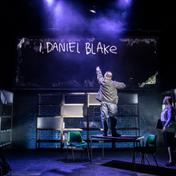 A man stands on a table facing a handwritten projection that reads 'I, DANIEL BLAKE', a woman stands on the stage next to him.