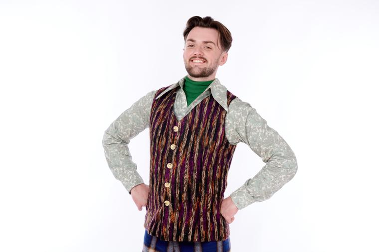 Max Guest with his hands on hips, smiling. He wears a purple and orange waistcoat with a floral shirt.
