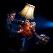 A man and a woman clutch onto an old lampshade as they lean forward. The woman wears an orange life-vest, a small spotlight points towards them.