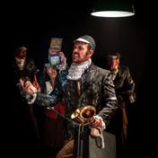 A man in a soiled black blazer grimaces, he holds a cigarette, and a cord that is attached to megaphone. He also holds a hard black briefcase. Three people stand behind him, partially hidden.