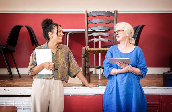 Two women smile at each other while leaning on a stage, one clutches a script while the other holds an iPad