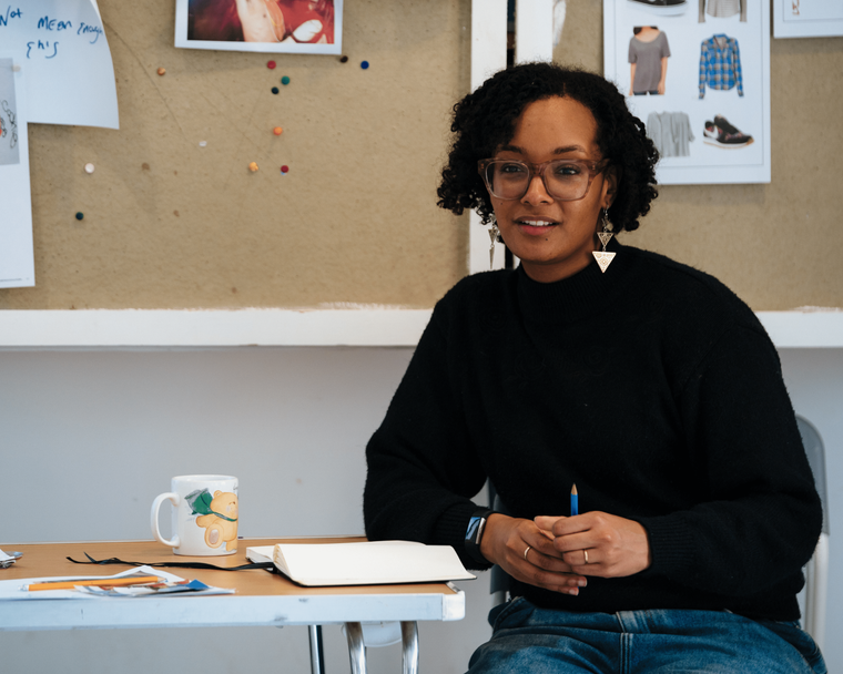A Black woman sat at a table holding a pen. She smiles, wearing glasses, a black jumper and blue jeans. She has short bobbed hair with dangly earrings showing.