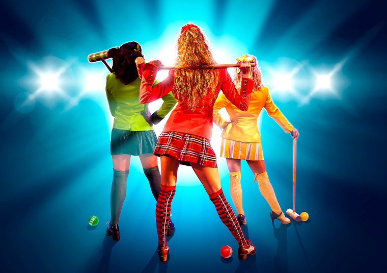 3 woman facing backwards holding wooden golf clubs, all dressed in different colours, wearing knee high socks, blazers and skirts. The background is blue with a line of stage lights shining forward.