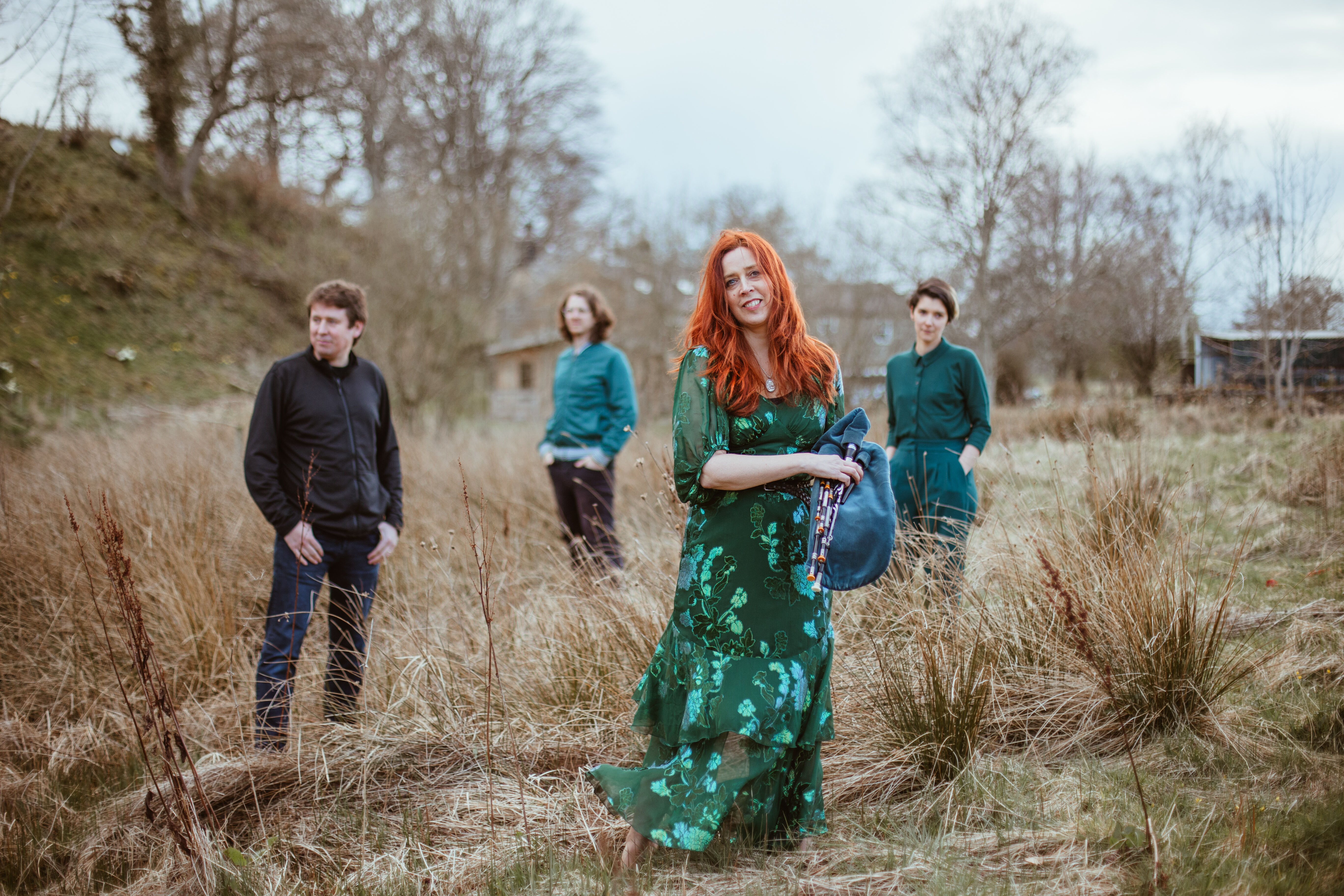 Image of Kathryn Tickell standing in a field wearing a long dark green dress with 3 other band member standing spaced out behind her looking in different directions