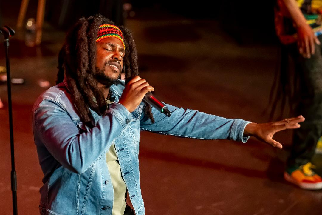 Man with dreadlocks and a head band singing into a hand-held microphone. His eyes are closed and he's reaching one arm out in front of him.