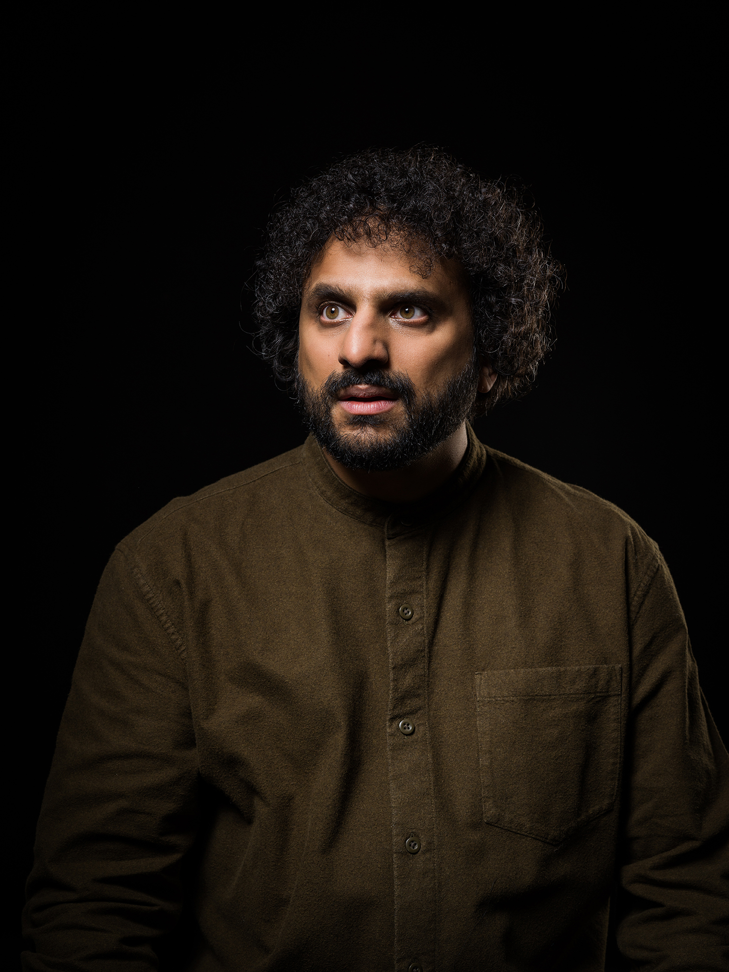 An image of Nish Kumar wearing a brown top, looking off to the side.