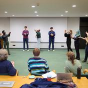 The cast of Two Gents stand in a square formation with their hands raised. Three people with their backs to the camera sit watching them.