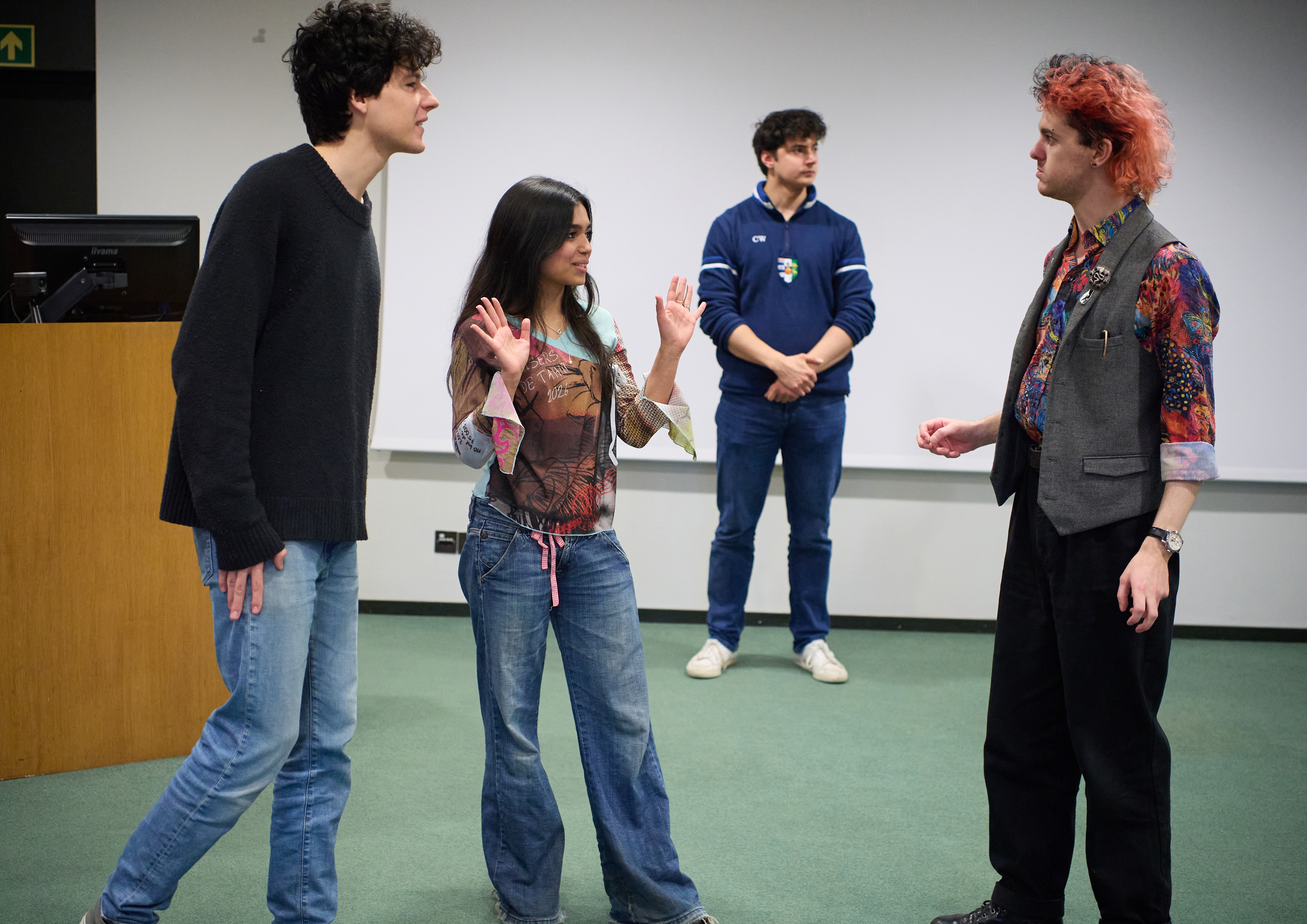 Three people stand in conversation in the foreground, another stands further away in the background. One of the three has their hands raised, while the other two stare at one another.