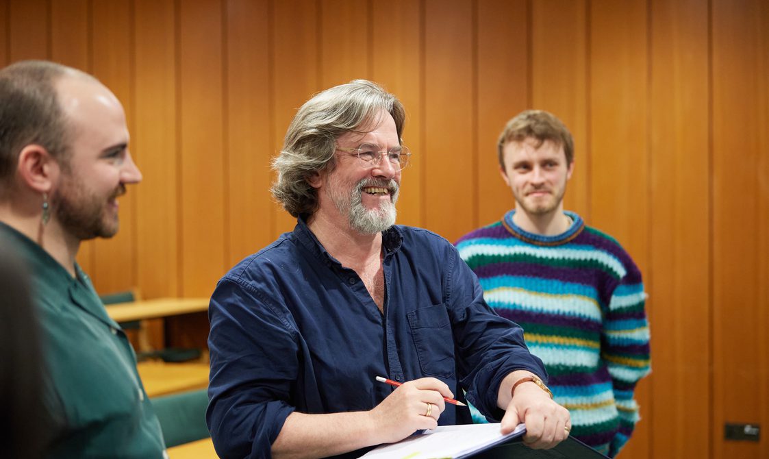Greg Doran smiles, holding a pencil above an open notebook. There are two other people either side of him smiling.