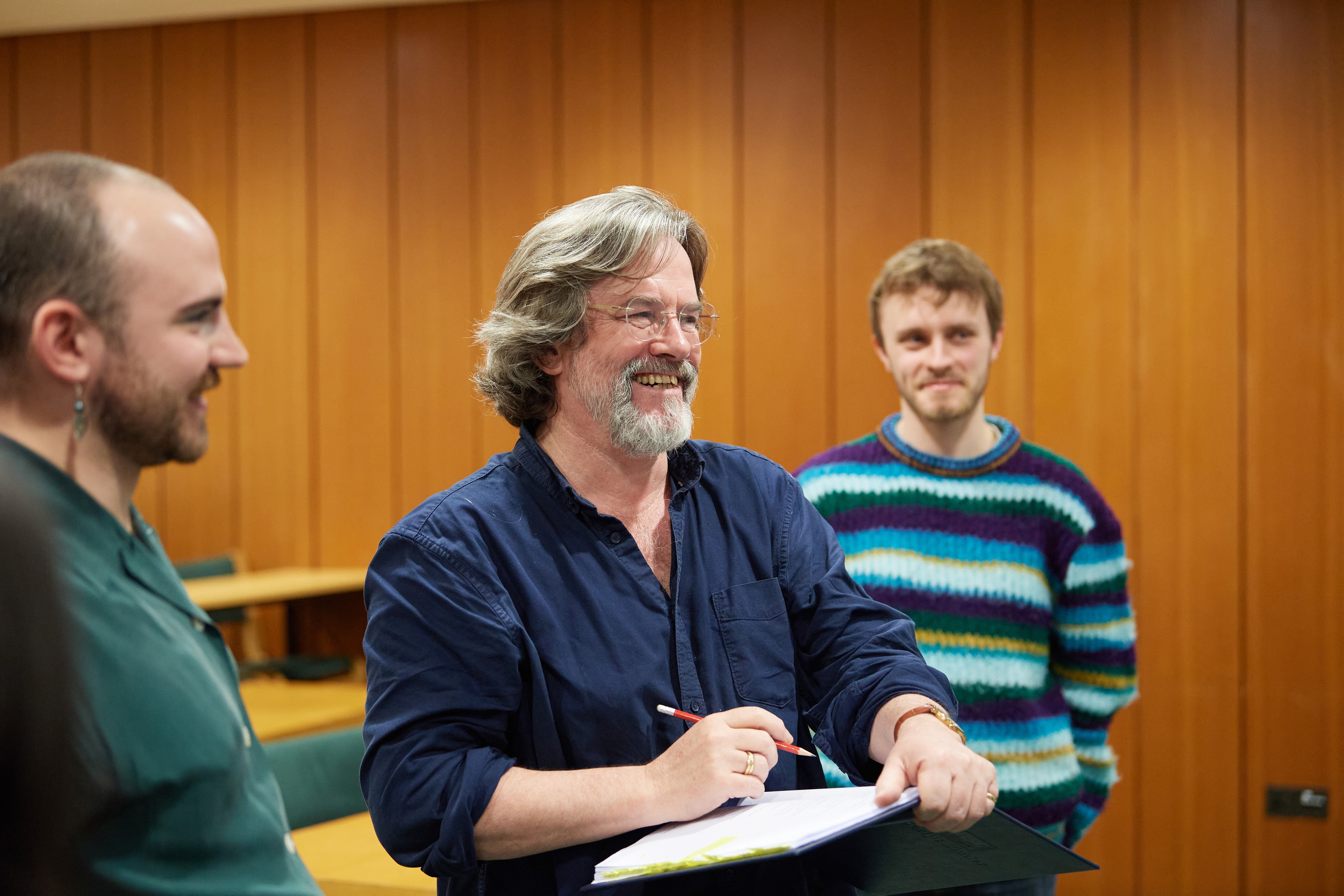 Greg Doran smiles, holding a pencil above an open notebook. There are two other people either side of him smiling.