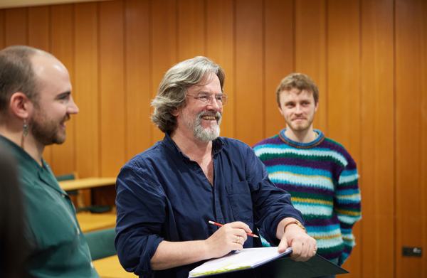 Greg Doran smiles, holding a pencil above an open notebook. There are two other people either side of him smiling.