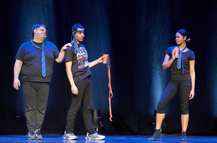 Three actors on stage. Two men stand closely together, one with his hand on the other's shoulder, a woman stands further away looking at them. Two of the actors wear loosened ties around their necks, while the other wears it around their head. Two of them hold party cups.