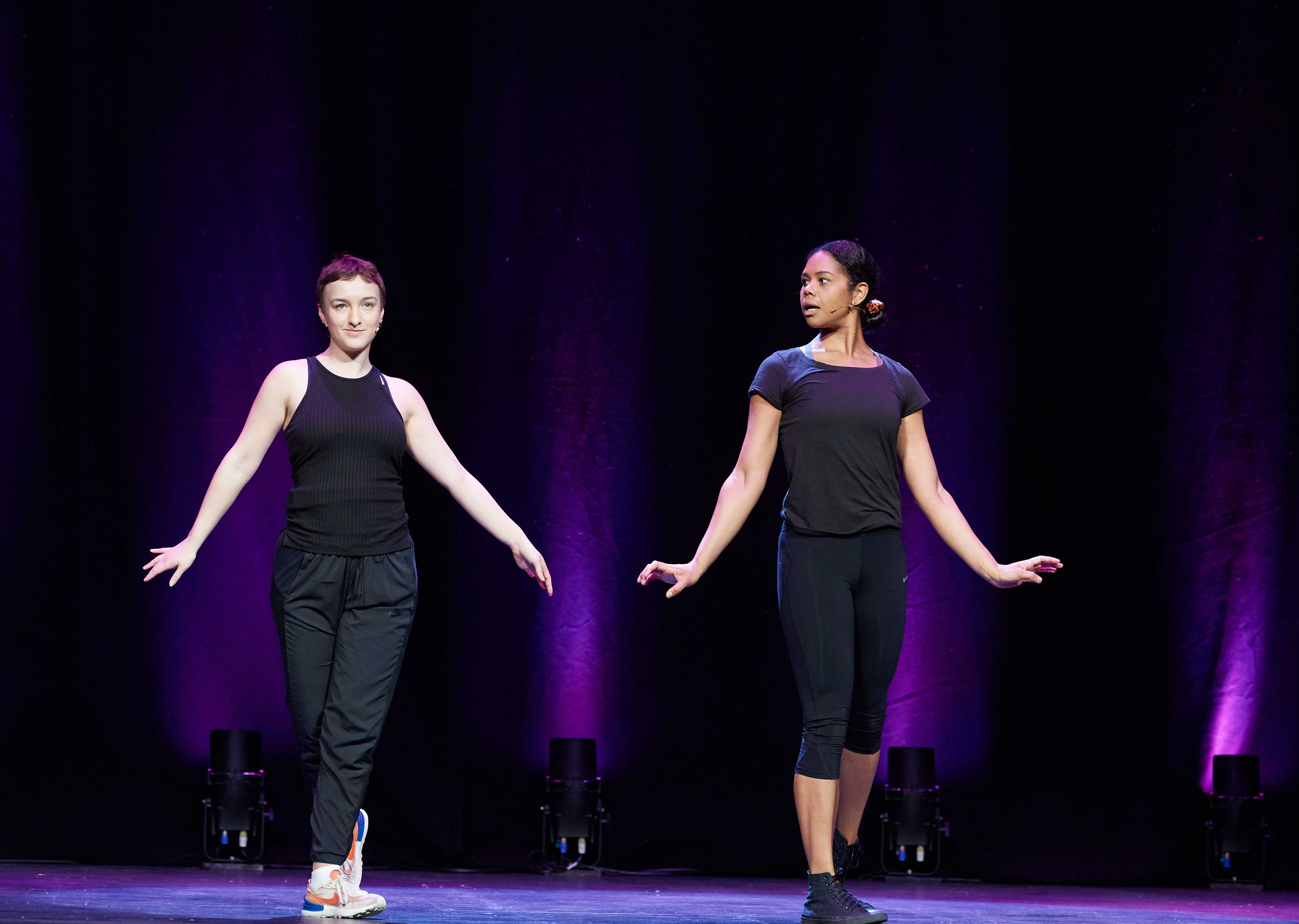 Two woman pose like dancers on stage. They stand next to each other with purple lighting behind them.
