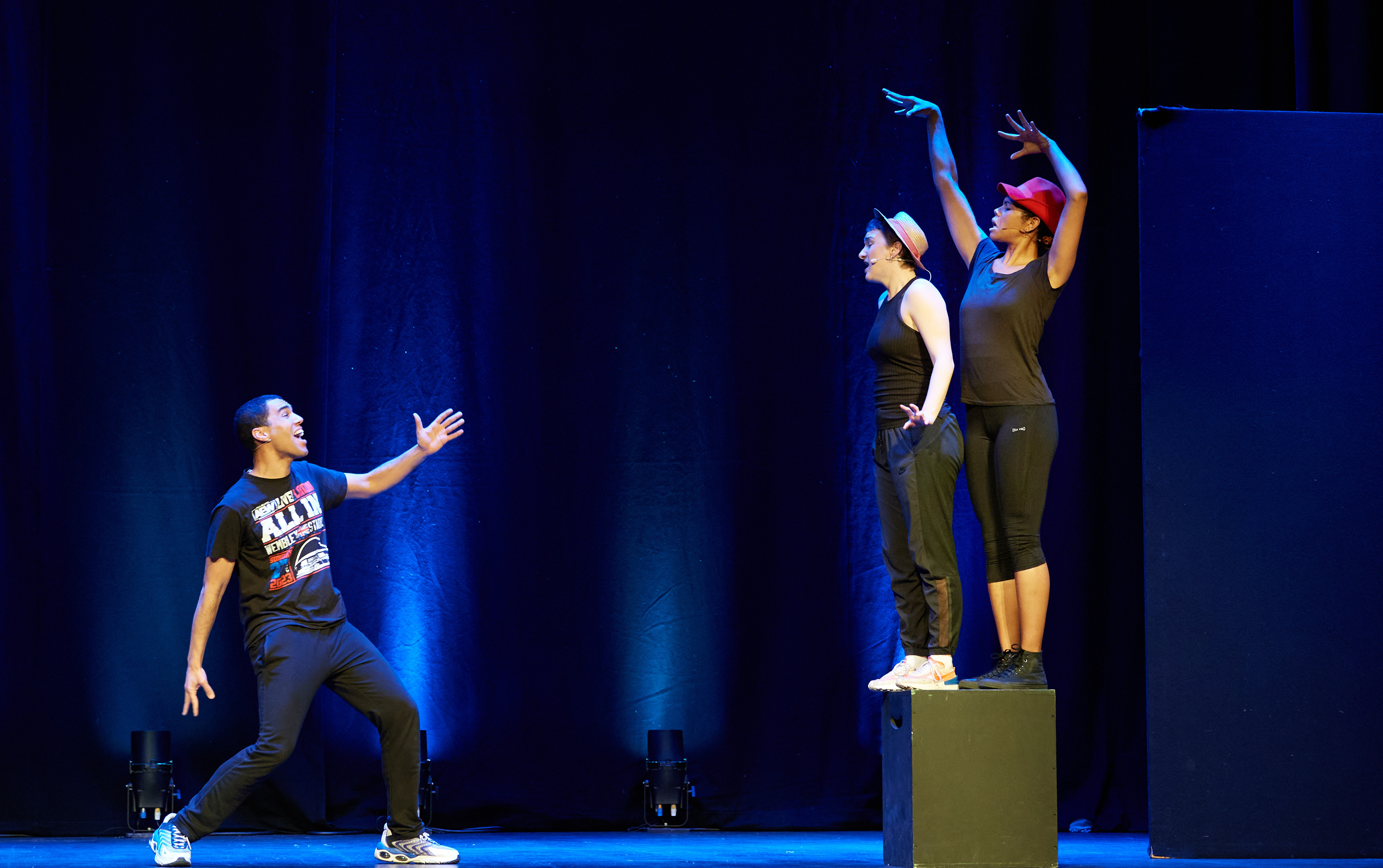 Two woman stand on a podium posing with hats on, a man stands below on the stage looking up with an arm outstretched