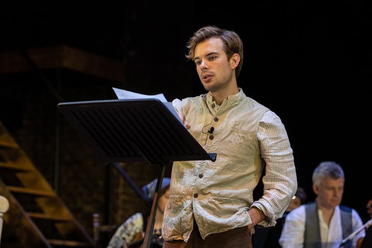 Charlie Hamblett stood behind a lectern on stage, he wears a beige waistcoat