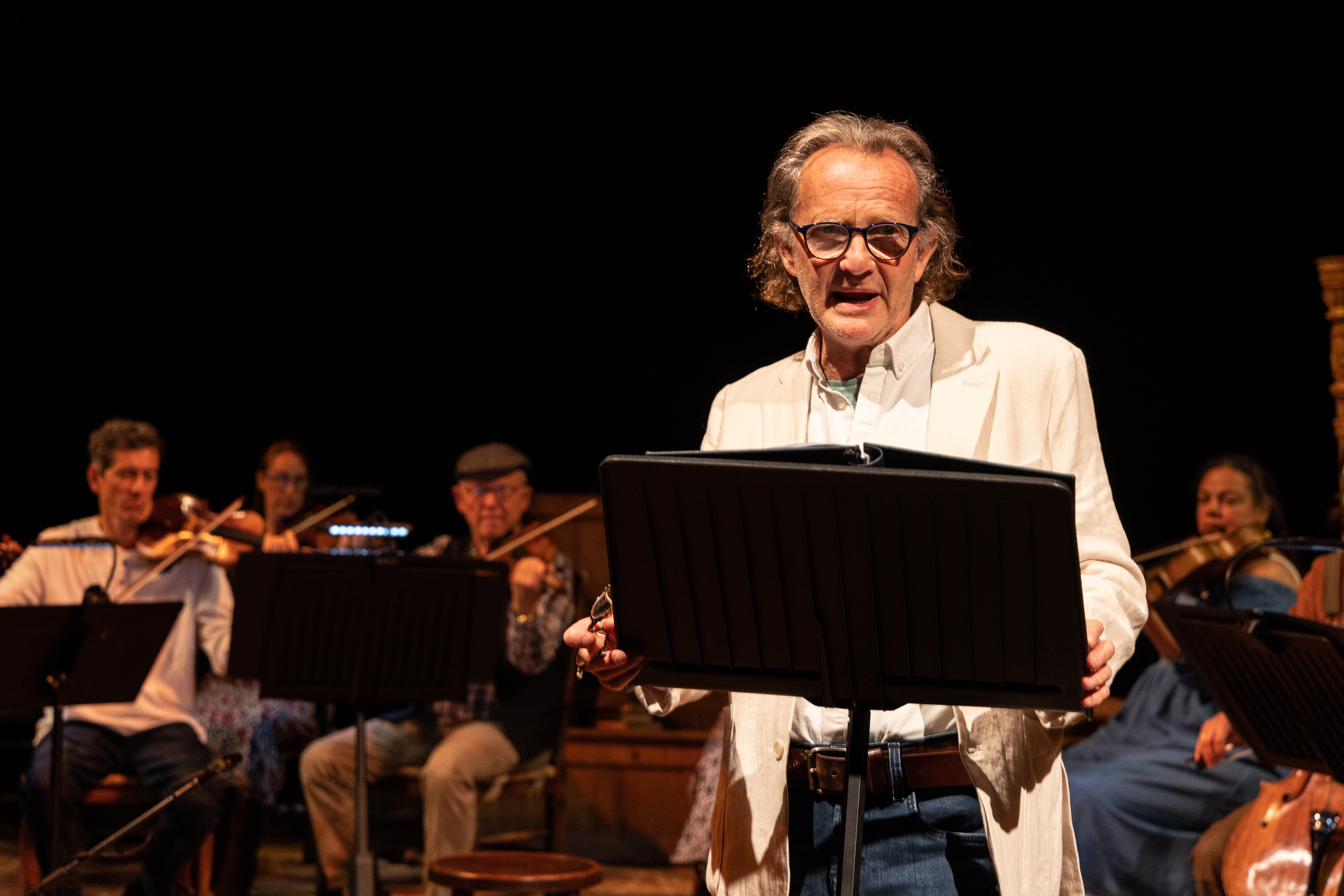 Anton Lesser stands behind a lectern, with Orchestra of the Swan playing instruments in the background
