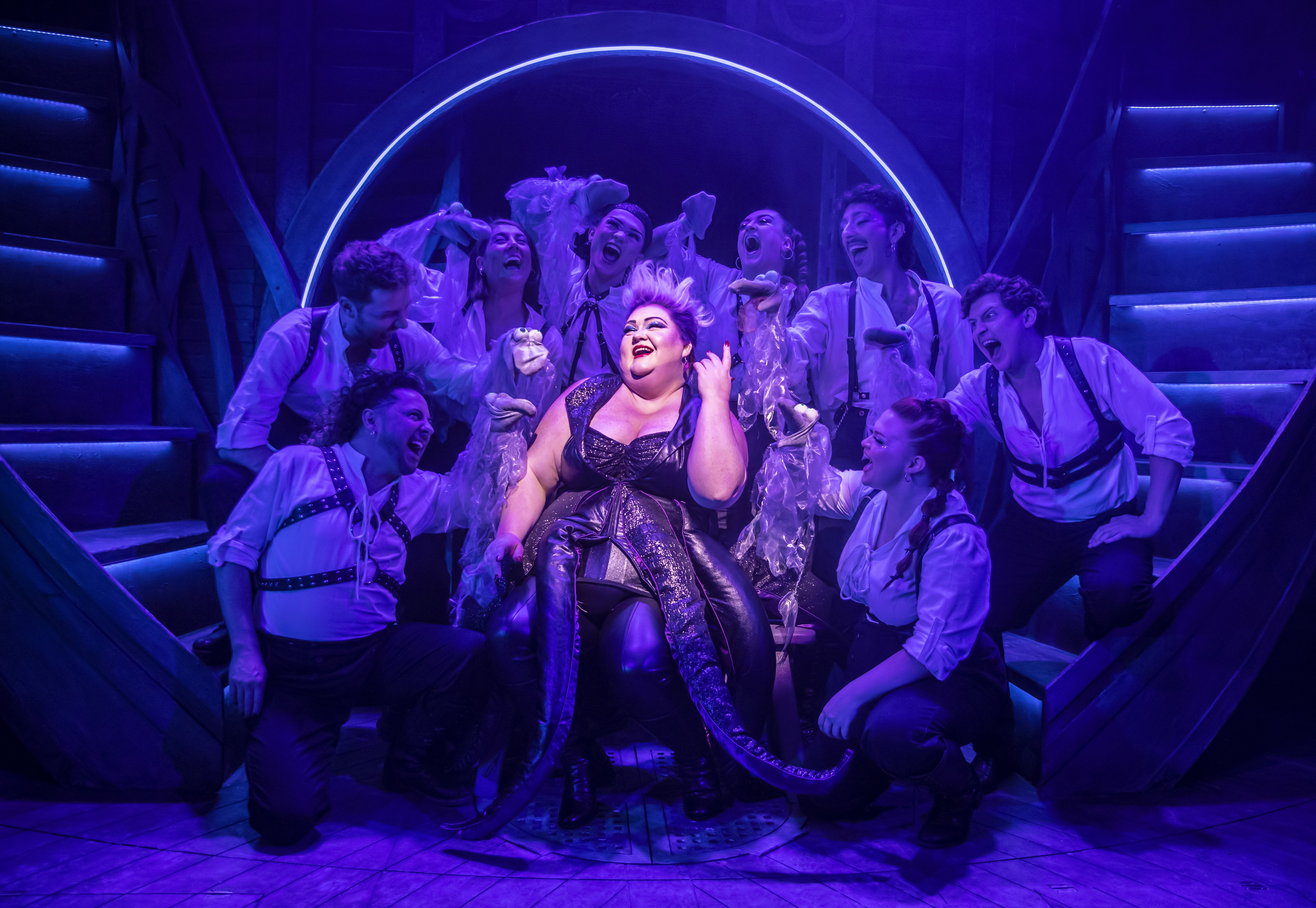 A photograph of the show Unfortunate. The character Ursula is lit up, bright against blue and purple lights, sat amidst an adoring crowd.