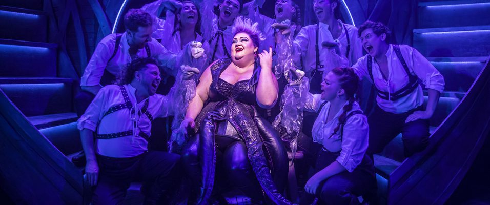 A photograph of the show Unfortunate. The character Ursula is lit up, bright against blue and purple lights, sat amidst an adoring crowd.