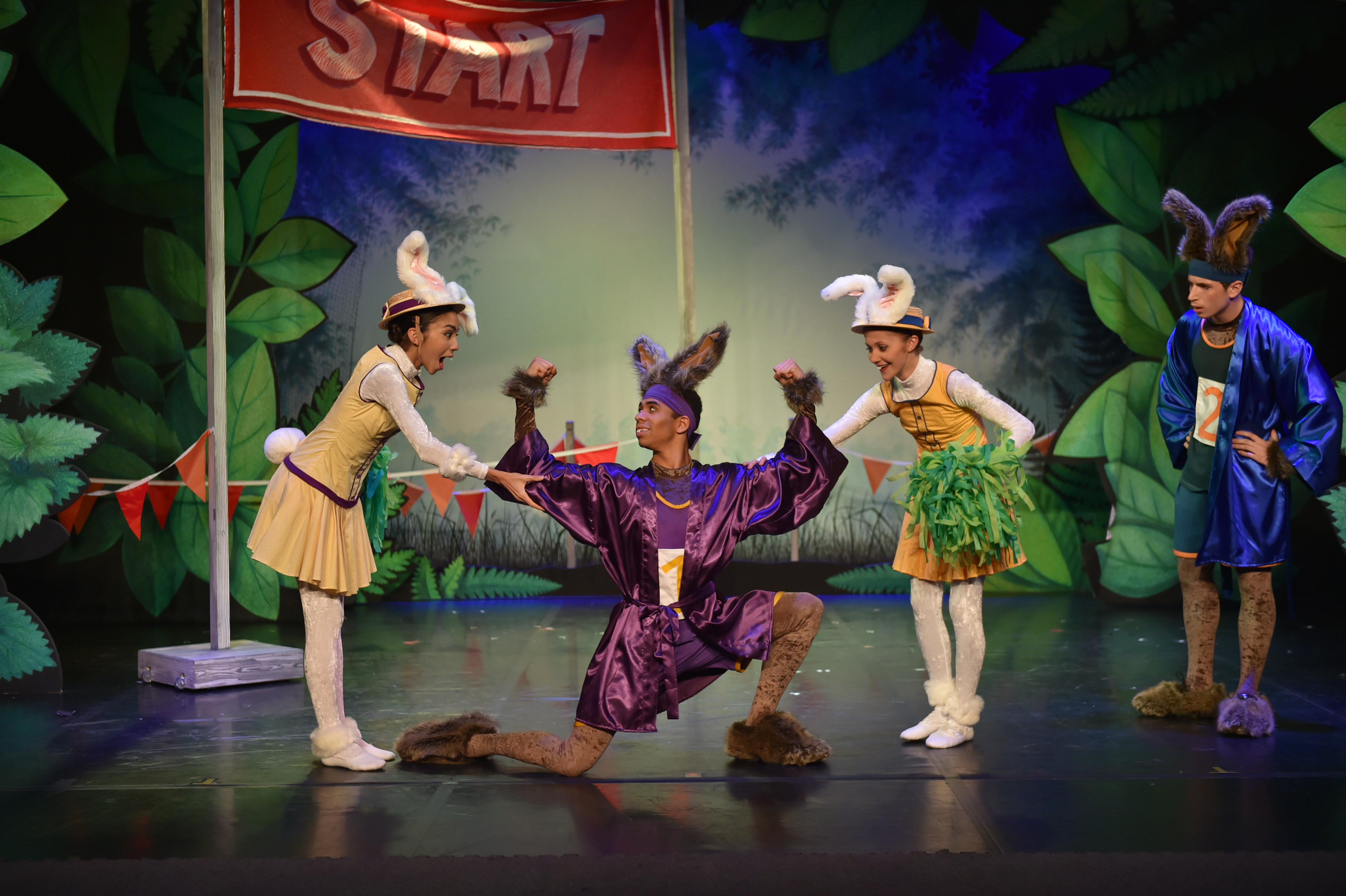 Four dancers on stage dressed as rabbits. One male dancer dressed in a purple gown is kneeling on one knee flexing his biceps and the two female dancers both wearing yellow dresses have one hand on each of his biceps. A male actor dressed in a blue gown is standing in the background with his hands on his hips.
