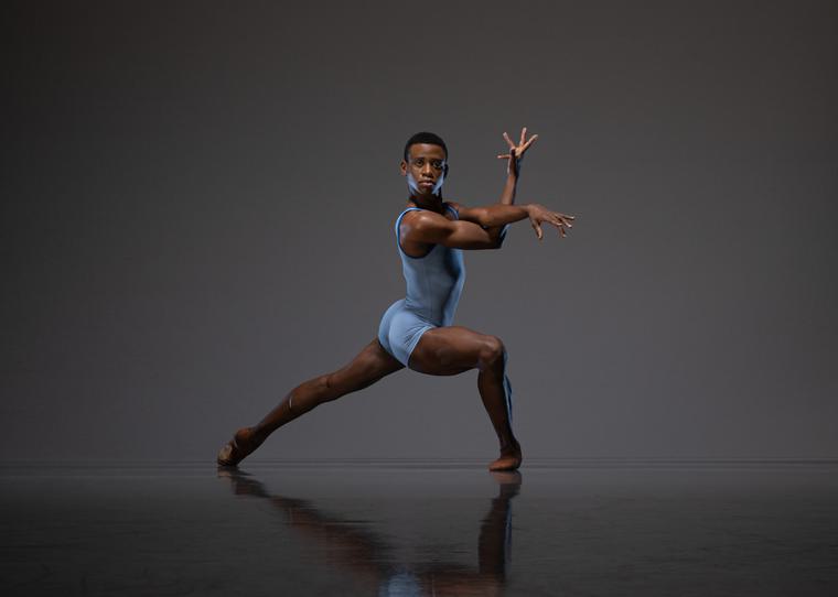 Acaoa de Castro performing contemporary dance onstage mid-move, leg extended backward, front leg bent, neutral expression, tension in arms which are extended and crossed, with fingers splayed