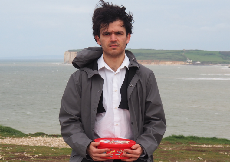 A man in a button up shirt with a rain coat sits along the sea with a box of Celebrations chocolates on his lap.