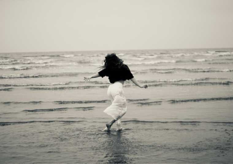 Black and white photograph of a woman dancing in the waves at the beach.