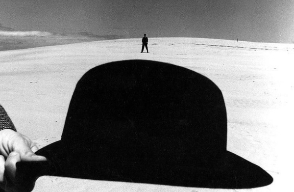 A black and white photograph of someone hold a bowler hat in front of a figure standing far away in a sandy dune landscape.