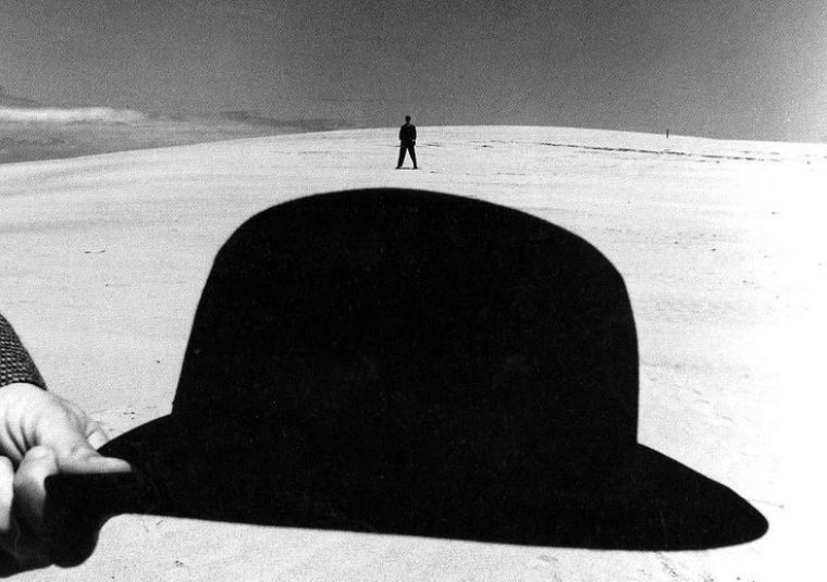 A black and white photograph of someone hold a bowler hat in front of a figure standing far away in a sandy dune landscape.