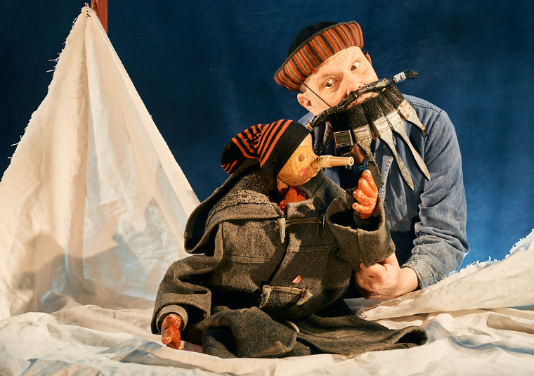 Actor on stage with in a whimsical costume of paint brushes for a beard. He is looking at a small carved doll, Pinocchio, who is dressed in an oversized jacket and beanie hat.