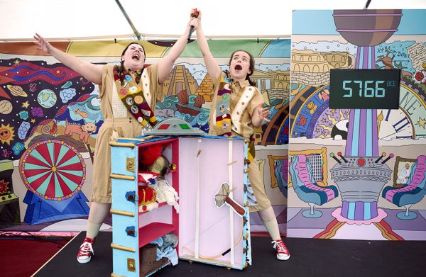 Two actors travelling through time fight over the gear shift and in mid yell, have broken and raise over their heads in despair. The stage is set with a colourful backdrop and a bright blue trunk with props spilling out.