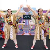Two actors dressed in brown jumpsuits with strips of different patterned fabric, sashes with various patches sewn on each, and bright red Converse shoes. Both actors are standing in the middle of the stage with a colourful backdrop, hands up in a star-like formation, smiling at the camera.