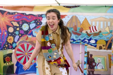 Actor singing towards the crown with her arms at her side. The backdrop is colourful illustrations of the beginning of the universe, historical depictions of great ships and ancient architecture including the pyramids.