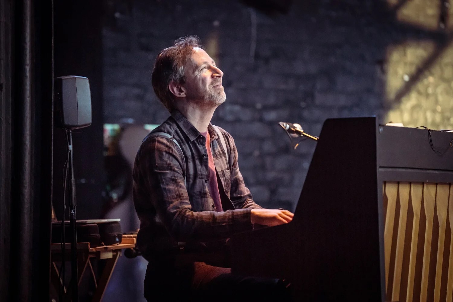 A man in a button down shirt sits at the keys smiling as he's playing on stage.