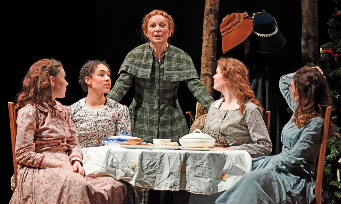 Four woman in floral dresses sit round a table. An older woman stands around them with her hands on the back of two of the chairs.