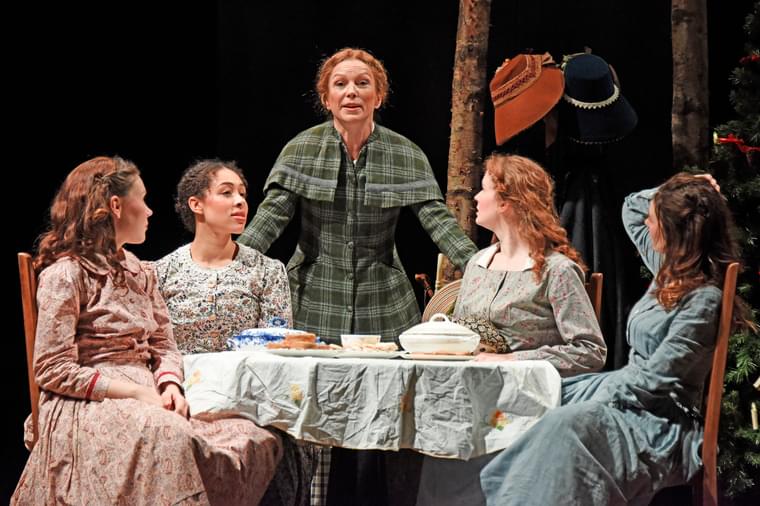 Four woman in floral dresses sit round a table. An older woman stands around them with her hands on the back of two of the chairs.