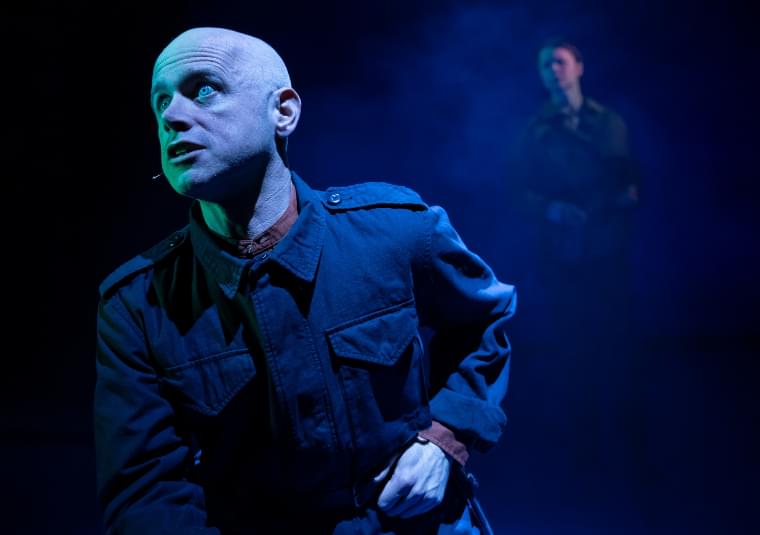 A man is kneeling down, side on, looking forward intently. In the background a figure can be seen standing behind within a cloud of smoke.