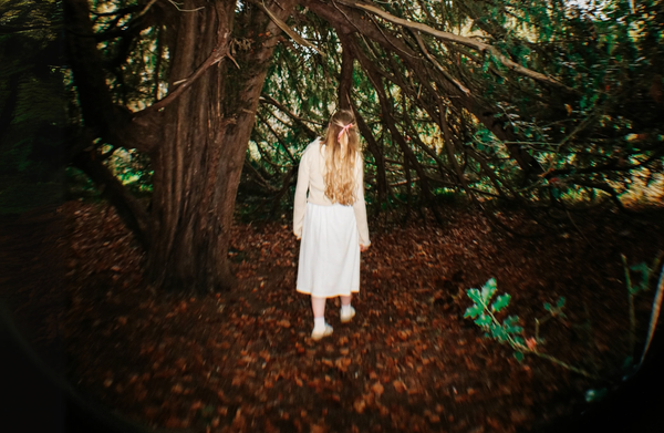 Taken with a fish eye lens, a figure in a white dress with long blonde hair adorned with a pink ribbon, walks deeper into the woods underneath a large tree. She walks away from the frame.