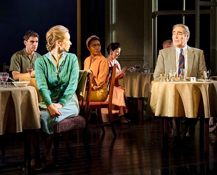 A man sat at a table looks over at a woman dressed in green at another table parallel, she turns her head to look at him. A couple at another table behind look over in the man's direction.