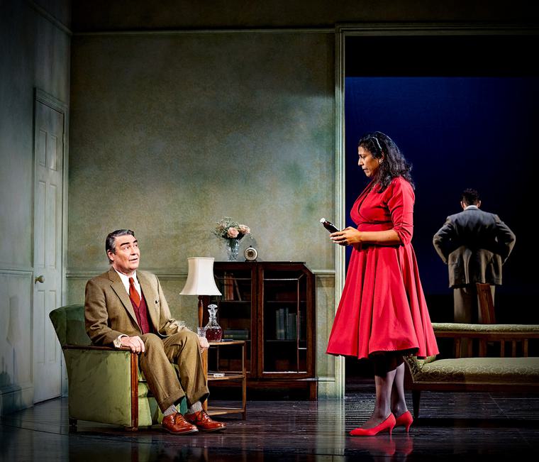 A man dressed in a brown suit sits in a green armchair. He looks up at a woman standing near him who is dressed in a bright red dress.