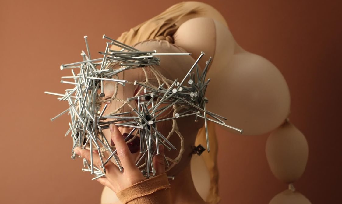 A person with dark red lipstick wears a mask made of tights, over their head, the tights are filled with balloons which gradually get smaller. They have a mask of nails covering their face.