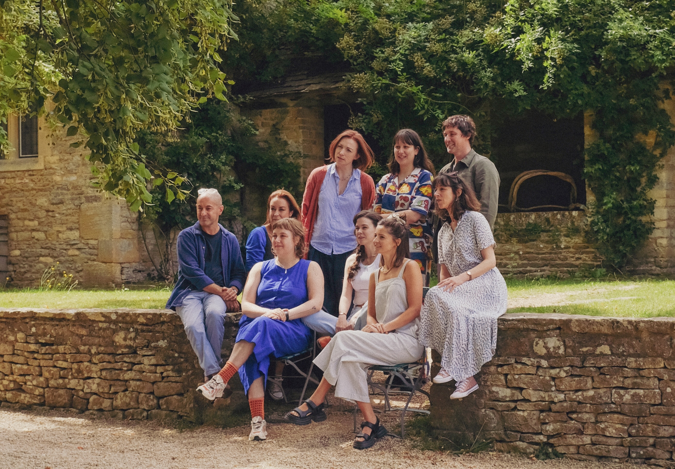 A group of people sit on a wall having their photo taken. There is a green lawn behind them and trees in front of a brick wall of a Manor House