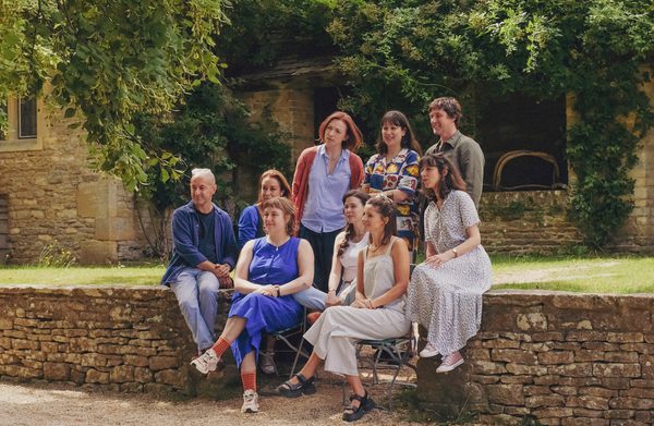 A group of people sit on a wall having their photo taken. There is a green lawn behind them and trees in front of a brick wall of a Manor House