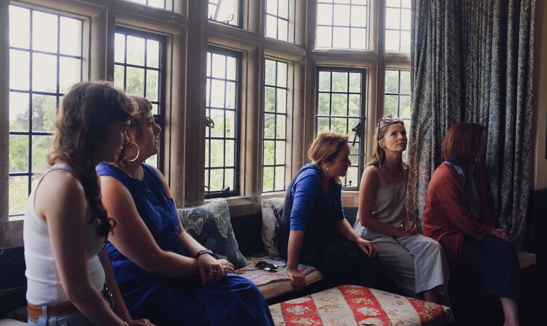 A group of people sit inside a Manor House on bench in front of a wall of glass windows