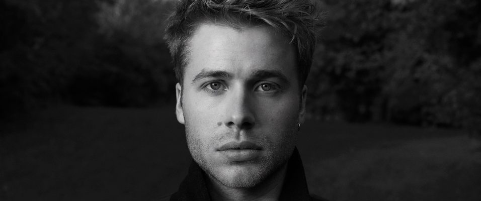 Black and white profile photograph of Ed Mcvey in a dark jacket with an atmospheric background of greenery and clouds.