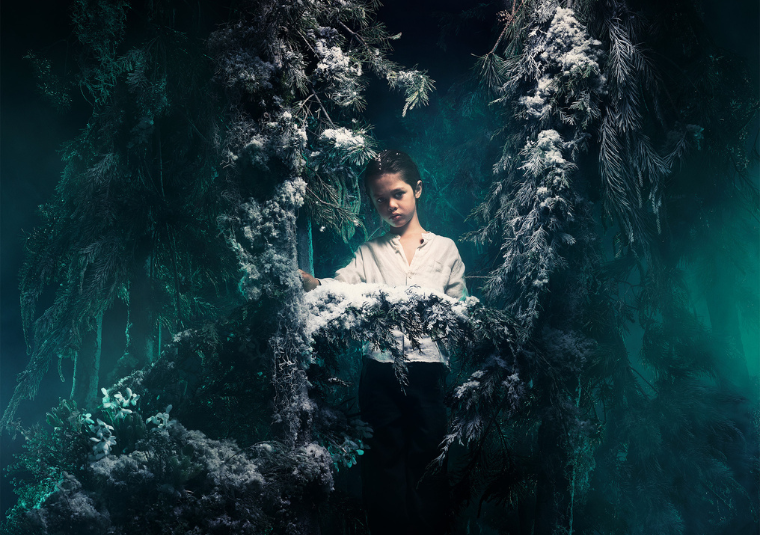 A child in a white linen shirt looking directly at the camera in the middle of a wintery forest with pines covered in snow.