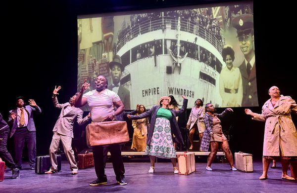On stage, people dance, old fashioned suitcases in hand or by their feet, behind them is projected the video of the Empire Windrush boat and people disembarking. Johnny is centre stage, holding his case, smiling with his hand raised, behind him people have their arms up, faces distressed.