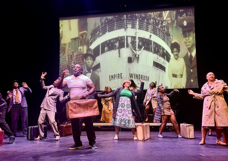 On stage, people dance, old fashioned suitcases in hand or by their feet, behind them is projected the video of the Empire Windrush boat and people disembarking. Johnny is centre stage, holding his case, smiling with his hand raised, behind him people have their arms up, faces distressed.