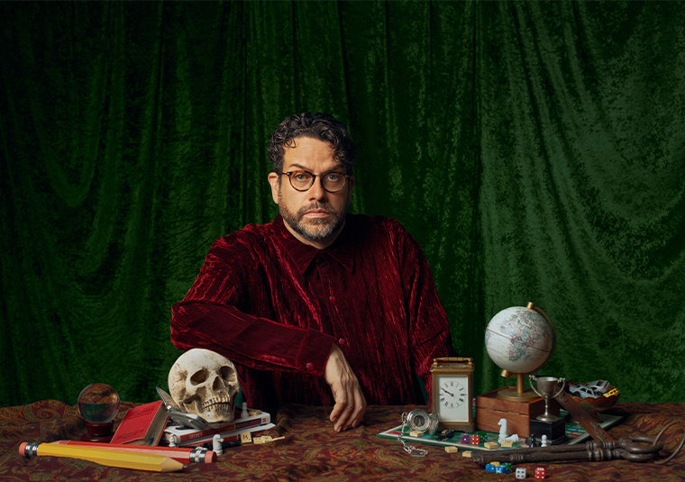 Michael Spicer is sat at a table in a red velvet jacket, face stern. His table is cluttered with things, including a skull, giant pencils, a small globe, a clock, dice, books, scrabble tiles