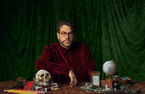Michael Spicer is sat at a table in a red velvet jacket, face stern. His table is cluttered with things, including a skull, giant pencils, a small globe, a clock, dice, books, scrabble tiles