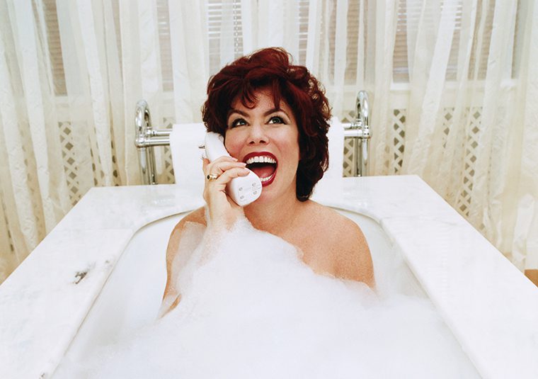Ruby Wax sits in a bubble bath, head back and mouth wide open, talking on a wireless landline phone.